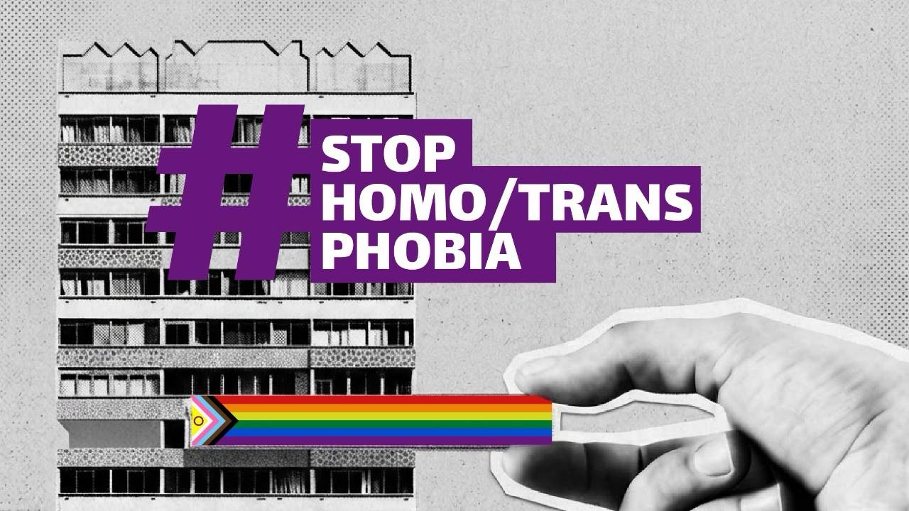 Black-and-white apartment building with a hand cutting through a rainbow Pride-colored strip using scissors. Purple text reads “# STOP HOMO/
