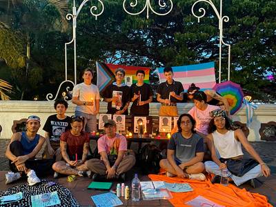 Grupo de personas con velas y banderas trans y LGBT+, reunidas en un altar con carteles y ofrendas al atardecer.